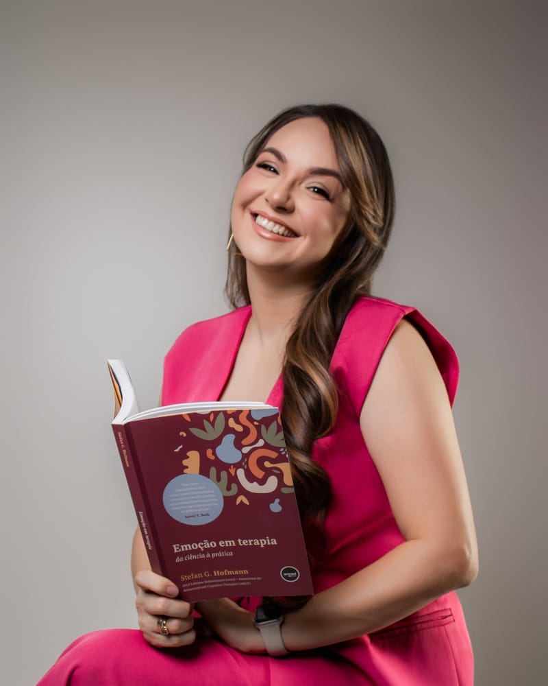 Juliani Pavão Fraga, psicóloga, sentada e sorrindo com um livro sobre emoções em terapia nas mãos